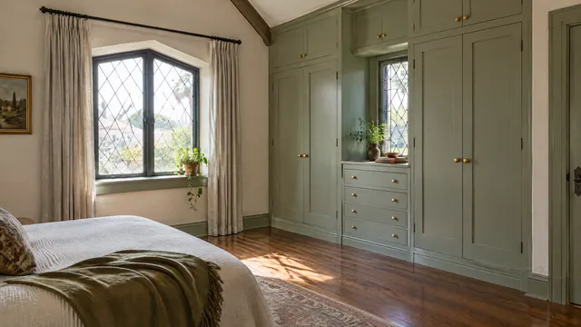 Bedroom in a 1920s historic Tudor-style home in the Willo district of Phoenix: built-in wardrobe cabinetry painted soft sage with brass knobs fitted beside a leaded-style window, original hardwood floors